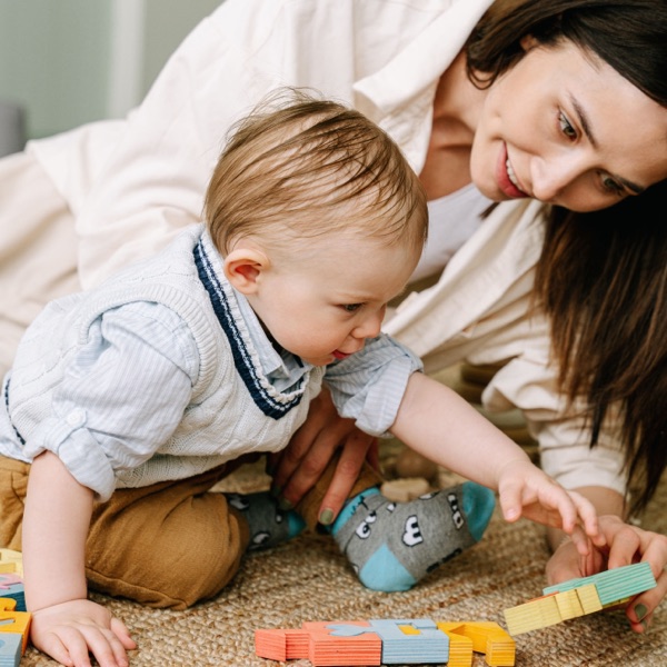 A parent lying on the floor beside her toddler, building with coloured wooden blocks